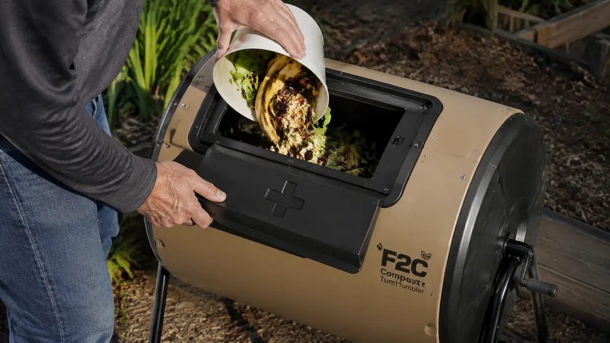 A hand sliding open the chamber door of a compost tumbler to add kitchen scraps